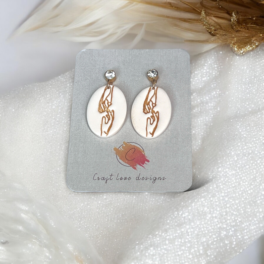 Elegant cream oval earrings featuring minimalist engraved hand illustrations in gold, displayed on a jewelry card over a soft, sparkling white fabric. 