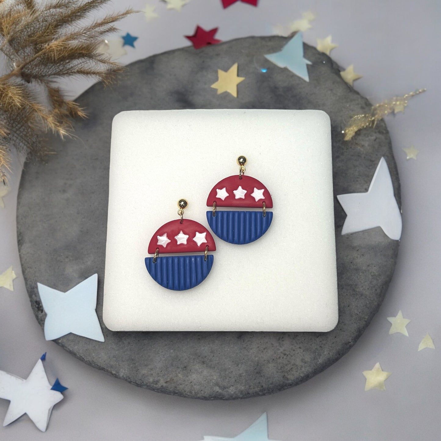 Festive red and blue 4th of July dangle earrings with white star details and gold hardware, resting on a neutral background.