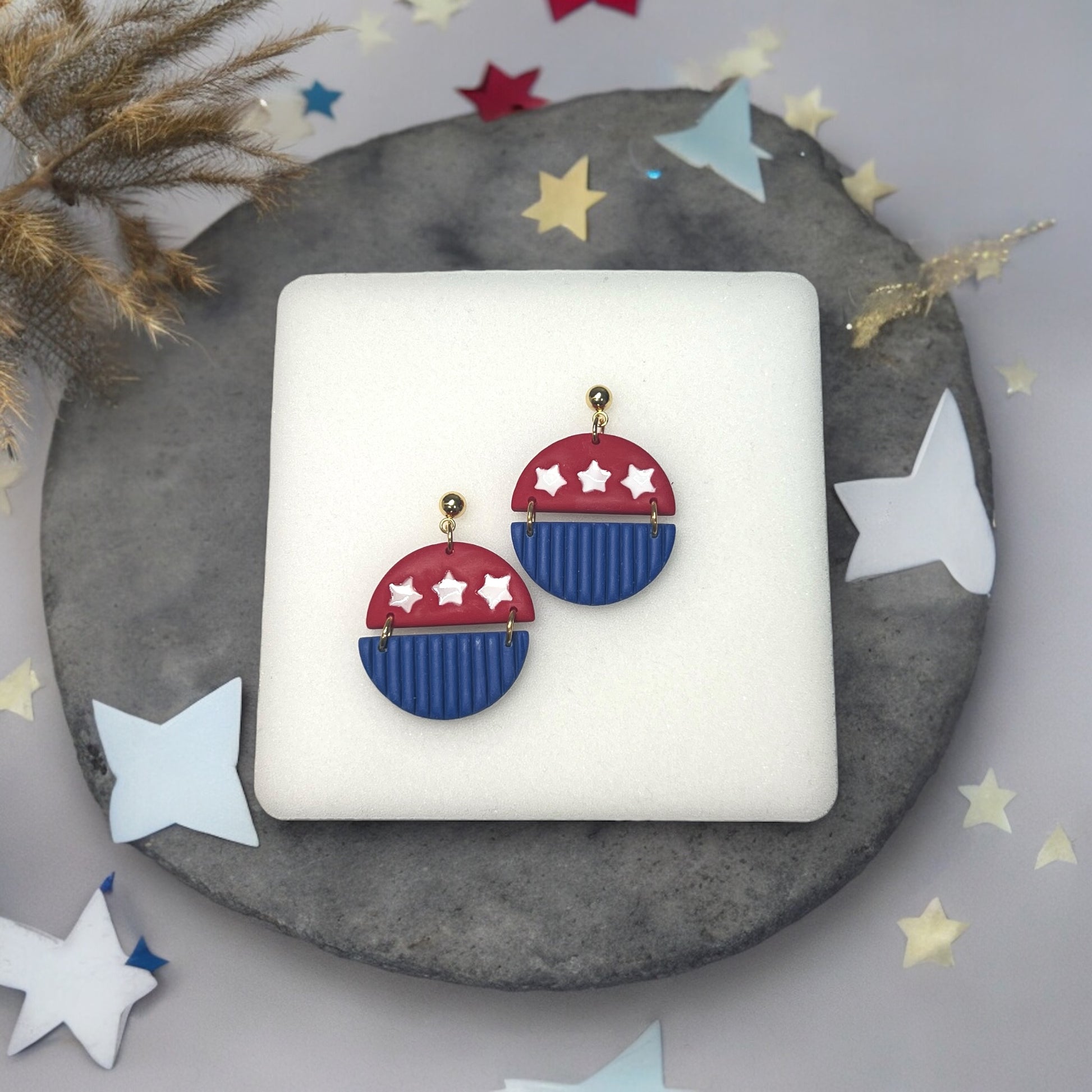 Festive red and blue 4th of July dangle earrings with white star details and gold hardware, resting on a neutral background.