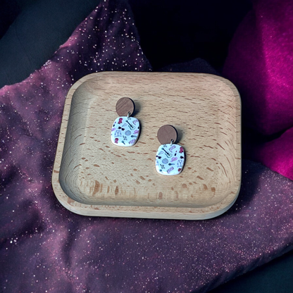 Oval drop earrings with wooden studs featuring fun, colorful patterns of guitars, hats, and jackets, placed in a wooden tray on cosmic fabric.