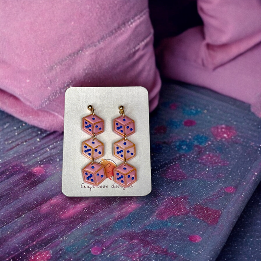 Long statement earrings featuring stacked translucent pink dice with blue dots, displayed on a branded card over a cosmic-patterned fabric. 