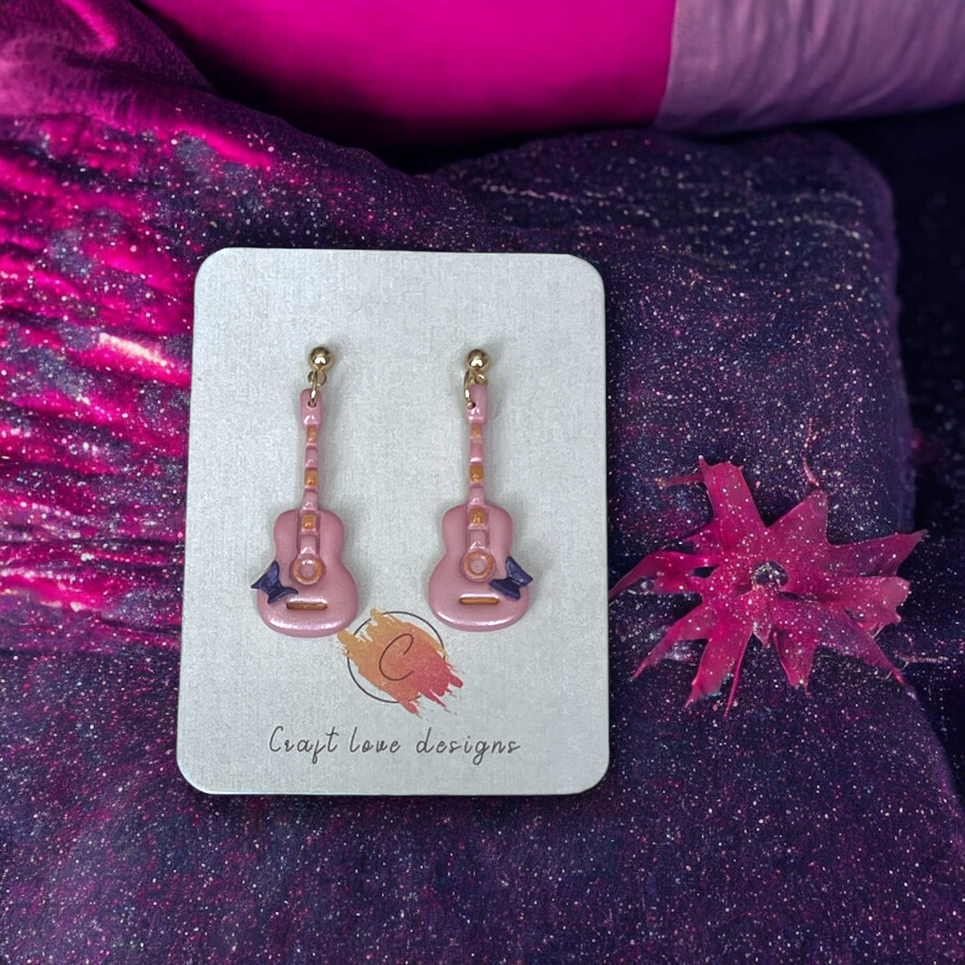 Handmade pink guitar-shaped earrings with gold accents and bow details, displayed on a Craft Love Designs card against glittery fabric.
