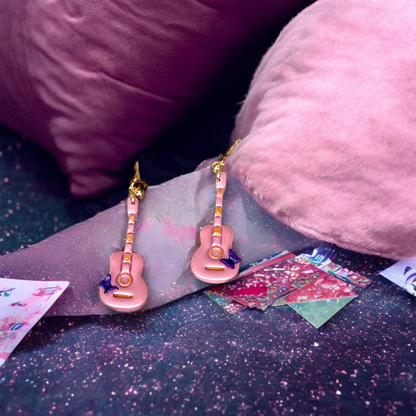 Pink guitar earrings with gold details and a bow resting on shimmering pink fabric, surrounded by decorative papers and cozy cushions.