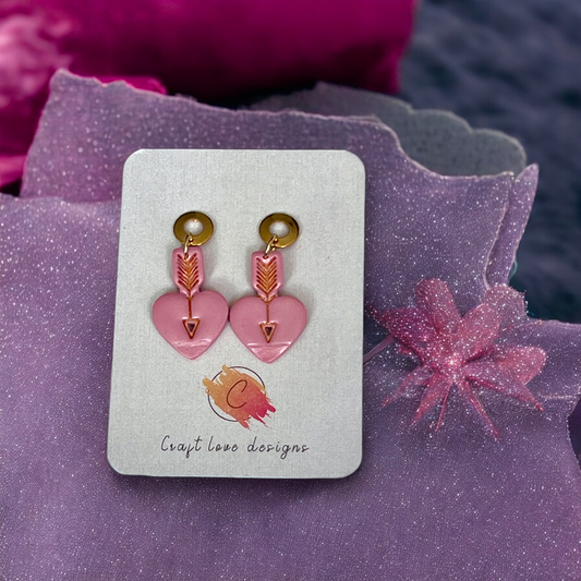 Handcrafted pink dangle earrings shaped as hearts with gold arrows running through, attached to gold accents, displayed on a branded card. 