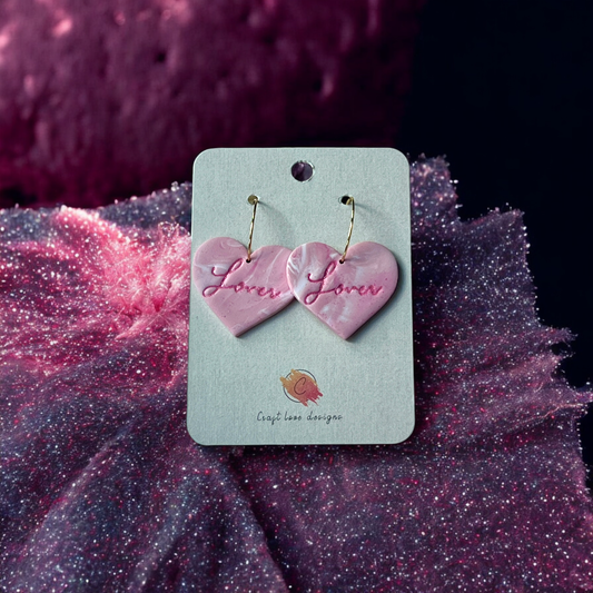 Pink heart-shaped dangle earrings with a marbled effect and cursive "Lover" engraving, displayed on a branded card against a sparkling fabric.
