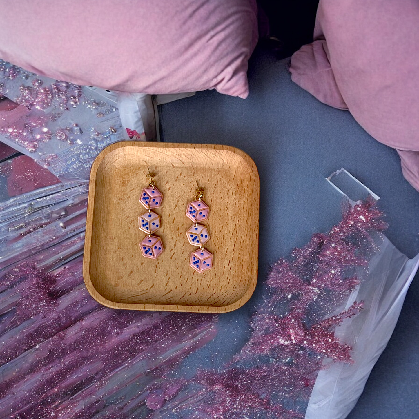 Pair of dangle earrings with stacked pink dice and blue dots, resting on a wooden tray surrounded by pink pillows and shimmering glitter decor.