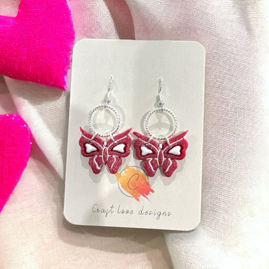 Pink butterfly earrings featuring white heart details, with a silver ring accent, perfect for a fun and stylish look.