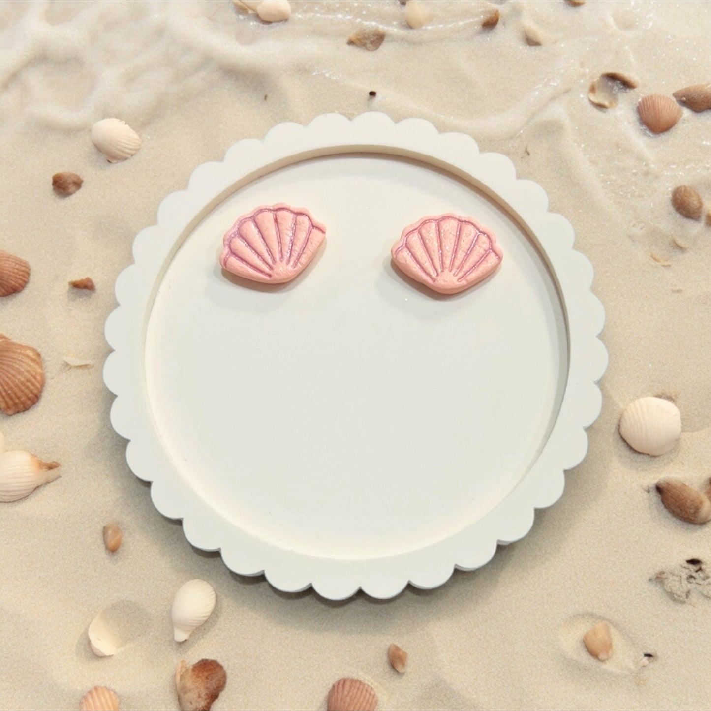 Light pink seashell stud earrings resting on a Craft Love Designs card, surrounded by beach shells and sand.