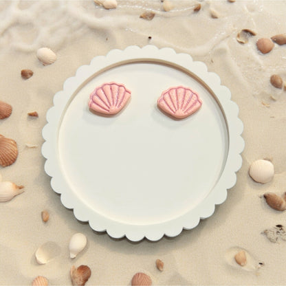 Light pink seashell stud earrings resting on a Craft Love Designs card, surrounded by beach shells and sand.