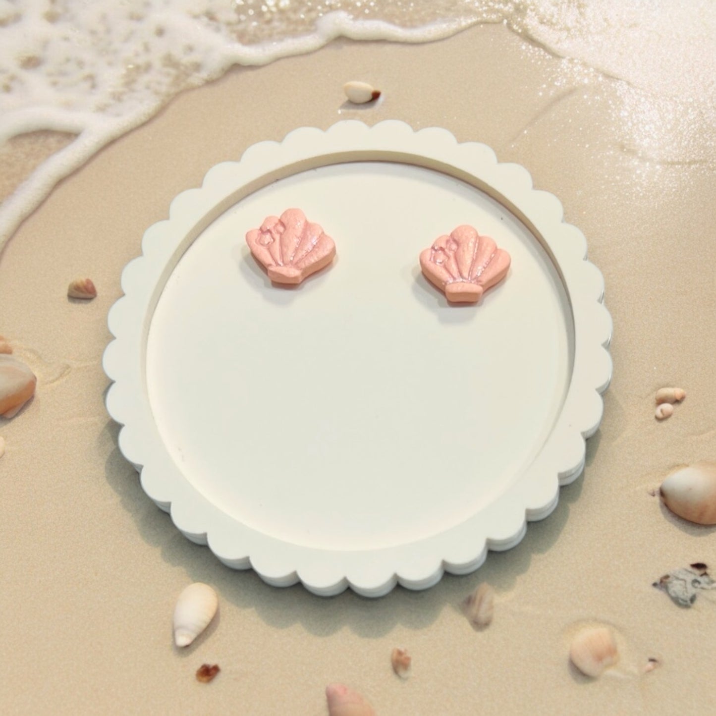 Close-up of handcrafted pink seashell earrings with a small bow accent, placed on a decorative white plate against a sandy beach background.