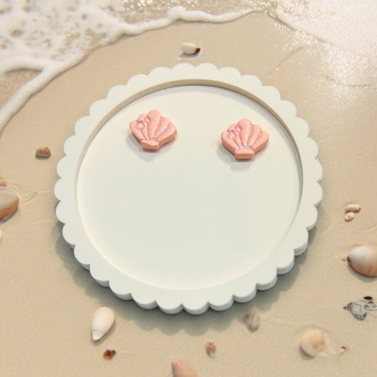 Close-up of handcrafted pink seashell earrings with a small bow accent, placed on a decorative white plate against a sandy beach background.