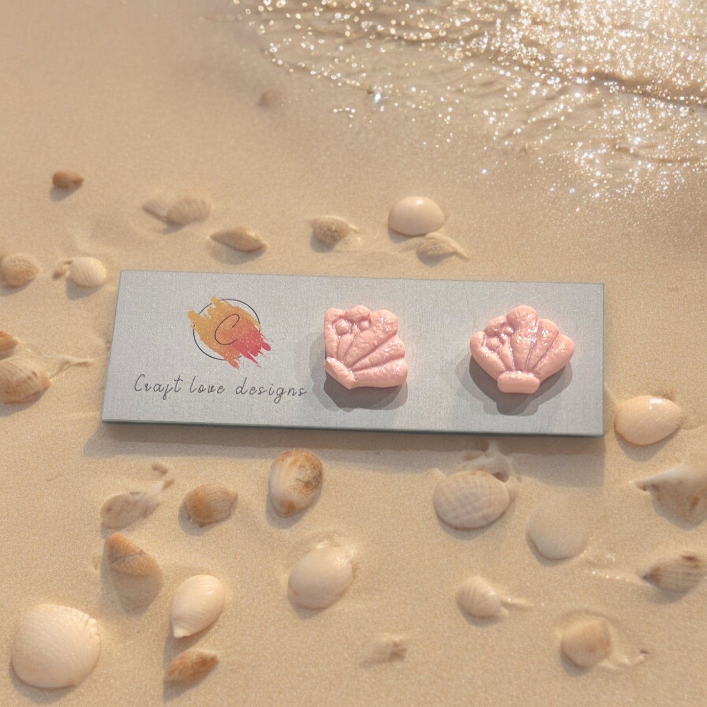 Pink seashell earrings with bow details, displayed on Craft Love Designs’ logo card, set amidst seashells on a sunlit beach. 