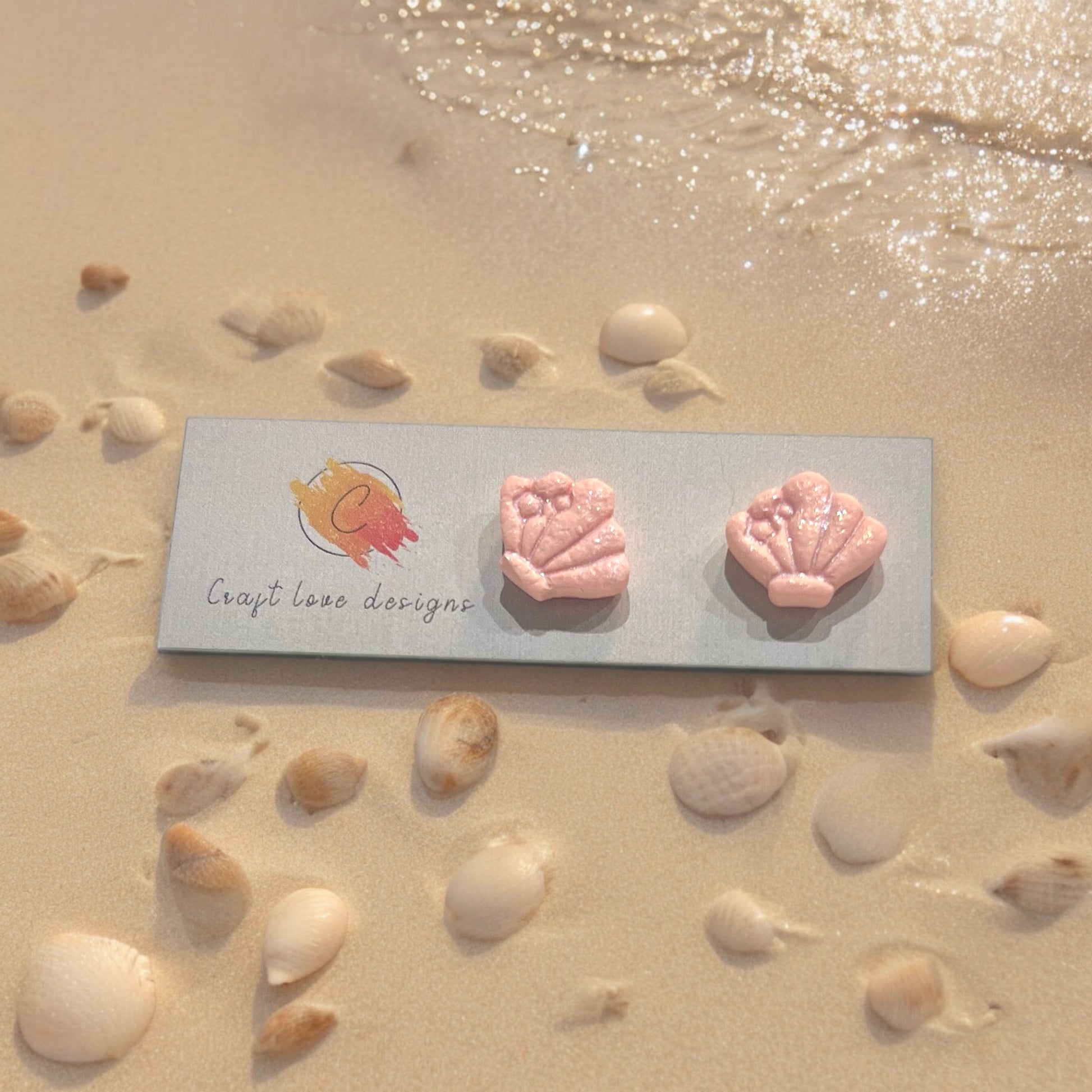 Pink seashell earrings with bow details, displayed on Craft Love Designs’ logo card, set amidst seashells on a sunlit beach. 