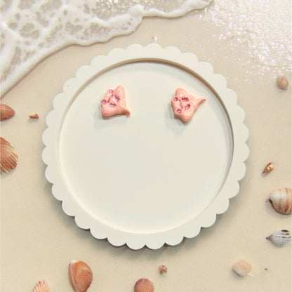 Pink stingray-shaped earrings displayed on a a round tray, lying on the sand with shells scattered around.