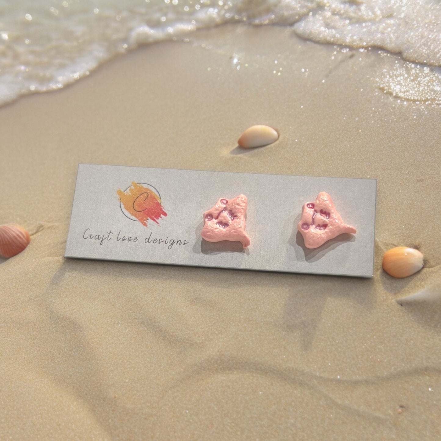 Pink stingray stud earrings resting on a decorative on a card with seashells and ocean elements in the background.