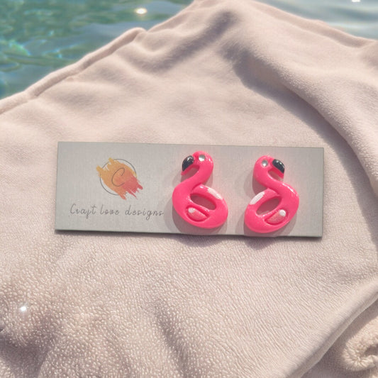 Close-up of pink flamingo earrings displayed on a jewelry card over a soft, cozy fabric near a pool setting. Fun summer accessory.
