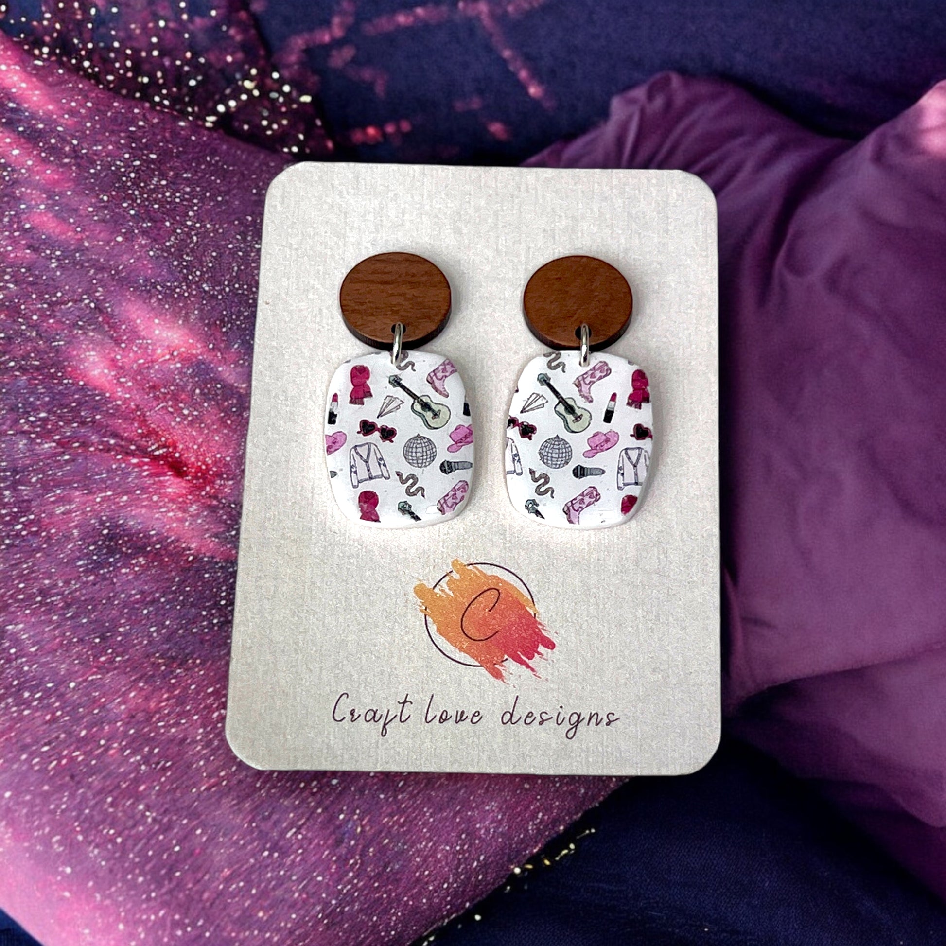Handmade earrings with printed patterns of guitars, jackets, and hats on white ovals, paired with wooden studs, displayed on a jewelry card. 