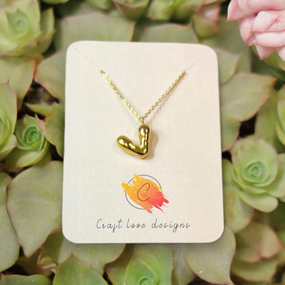 Petite puffy V heart pendant necklace displayed on a branded card, with golden highlights surrounded by vibrant succulents and soft light.