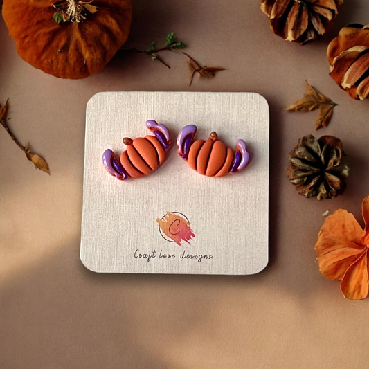 Festive pumpkin-shaped earrings with vibrant orange tones and purple accents, displayed on a card surrounded by autumn leaves and decor.