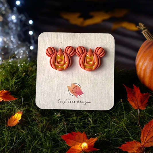 Festive pumpkin-shaped stud earrings with Mickey-inspired ears, displayed on a branded card over a grassy surface with fall leaves and lights.