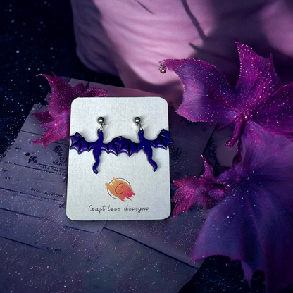 Purple bat-shaped earrings with silver studs displayed on a Craft Love Designs card, set against a pink and purple dreamy background.