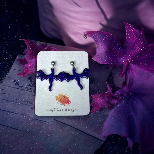 Purple bat-shaped earrings with silver studs displayed on a Craft Love Designs card, set against a pink and purple dreamy background.
