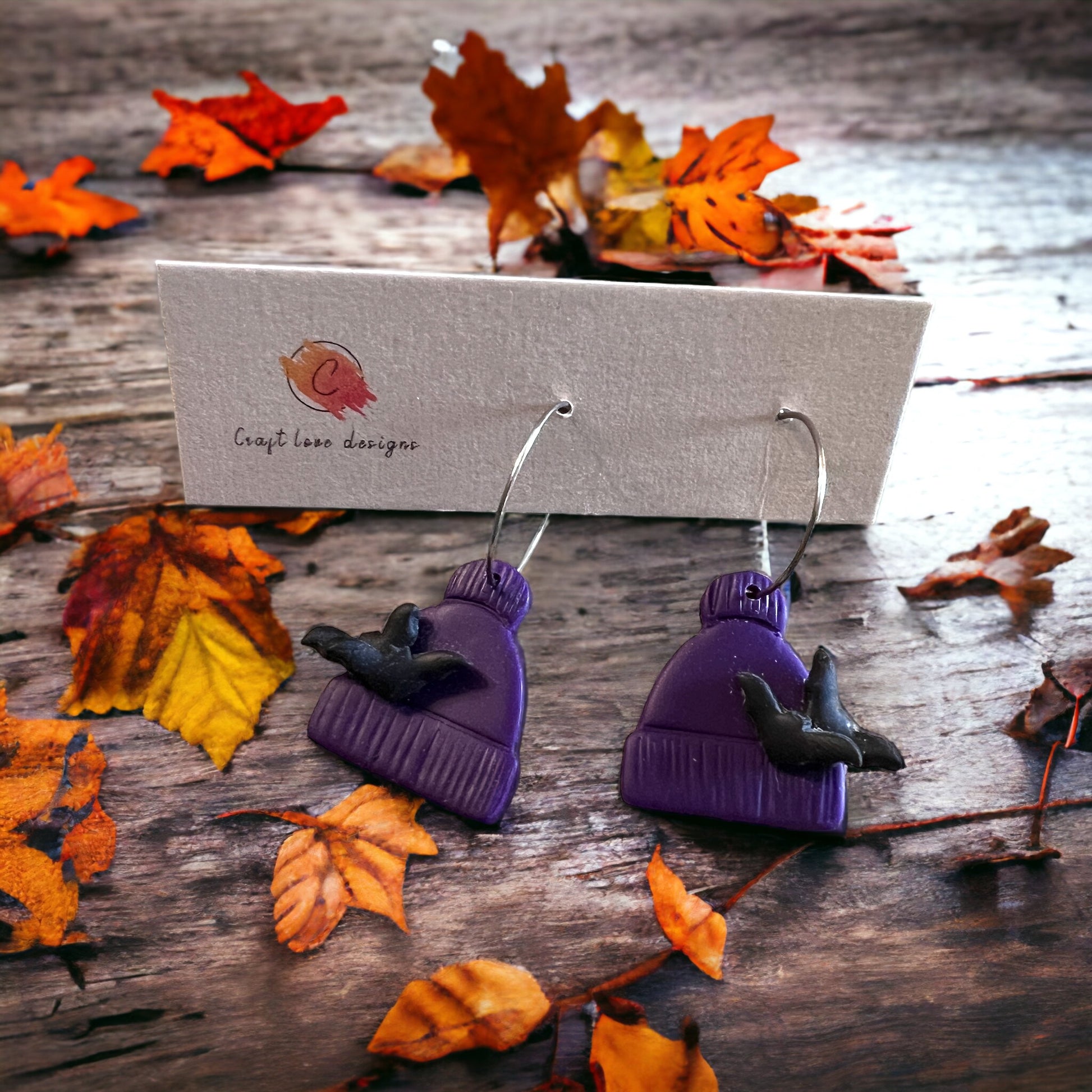 Purple beanie-shaped polymer clay earrings featuring a small bat design. Fall-themed dangles perfect for seasonal wear. 