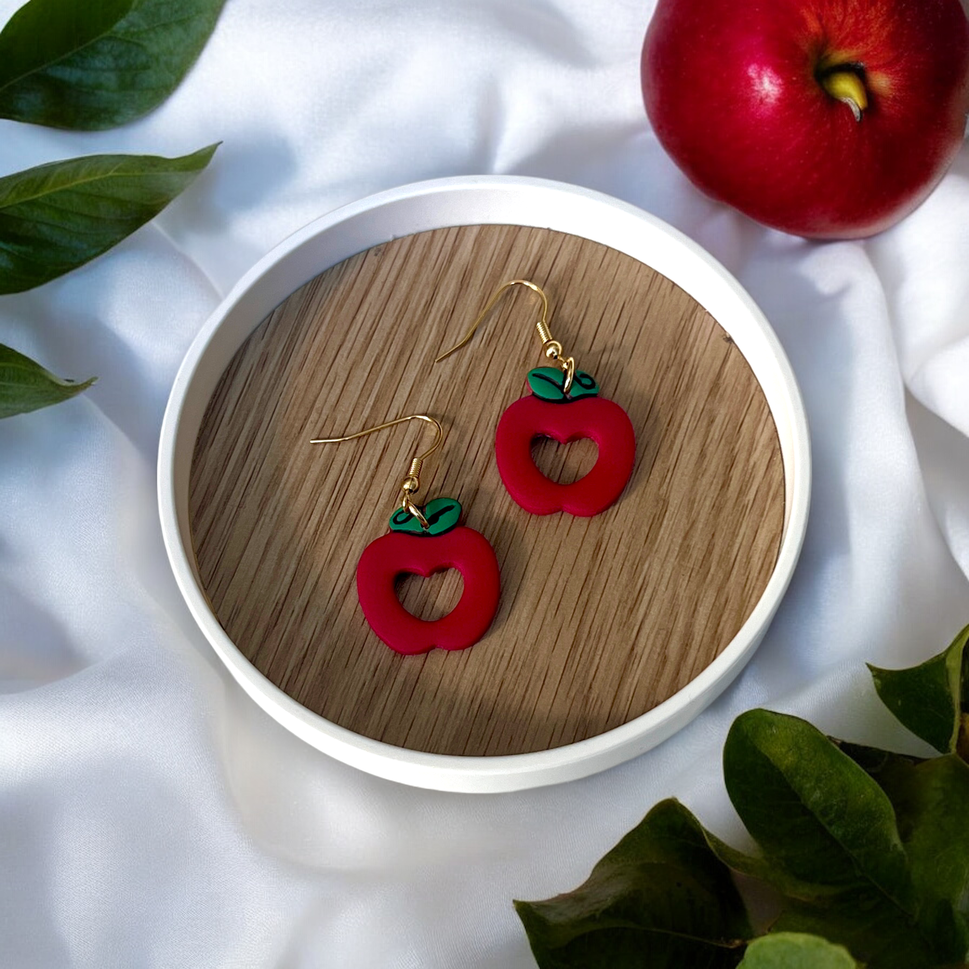 Fun red apple earrings with green leaves displayed on a wooden tray, ideal for adding a fresh pop of color to your style.