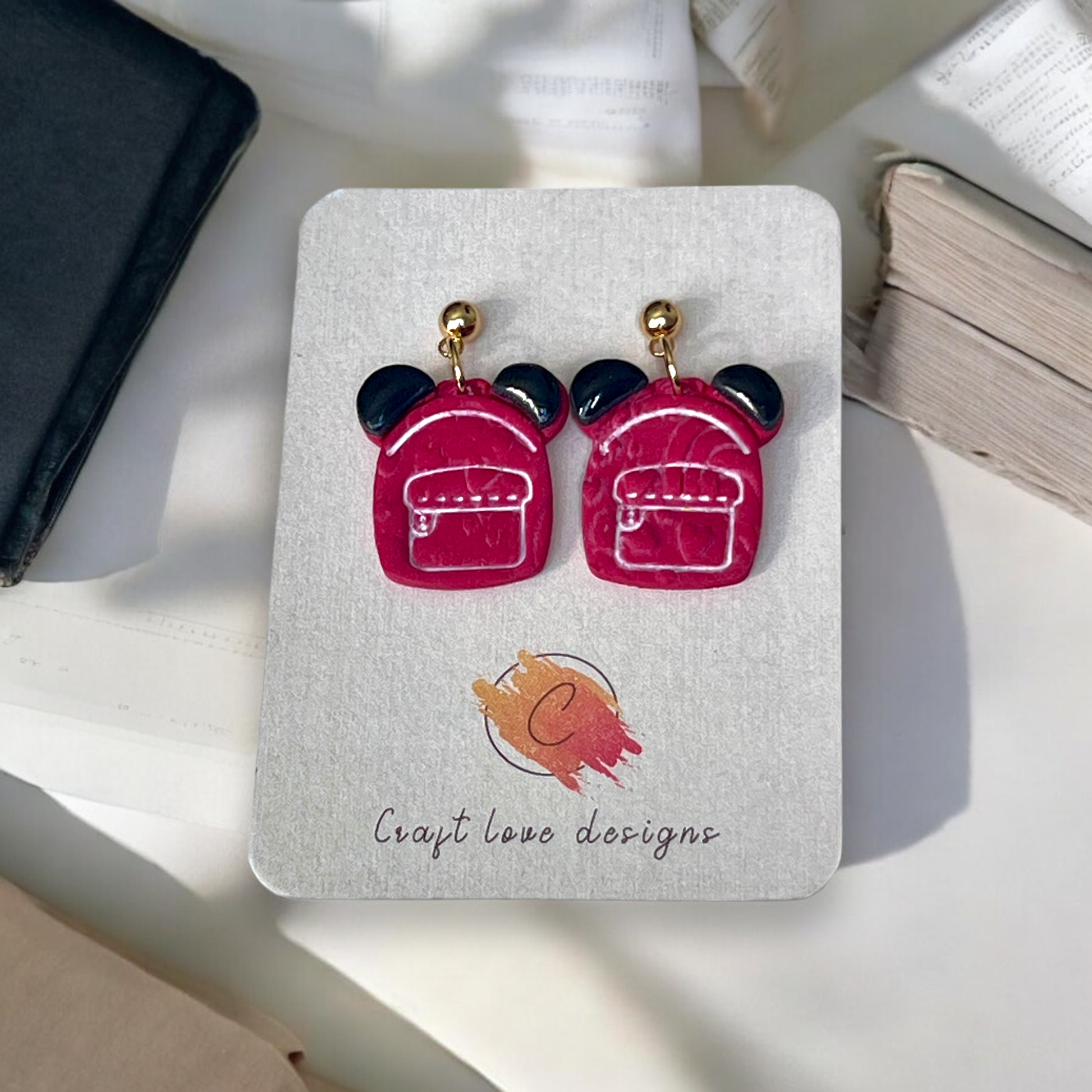 Playful handmade red backpack drop earrings with black ear accents and gold stud tops, displayed on a designer card over a soft fabric backdrop.