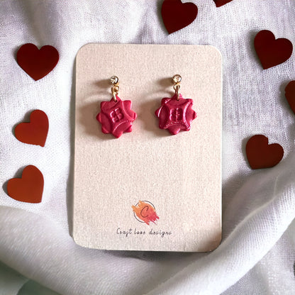 A close-up of red cubic zirconia dainty dangle earrings on a beige display card, surrounded by red heart decorations for a romantic vibe.