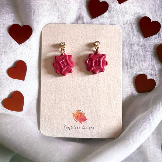 A close-up of red cubic zirconia dainty dangle earrings on a beige display card, surrounded by red heart decorations for a romantic vibe.