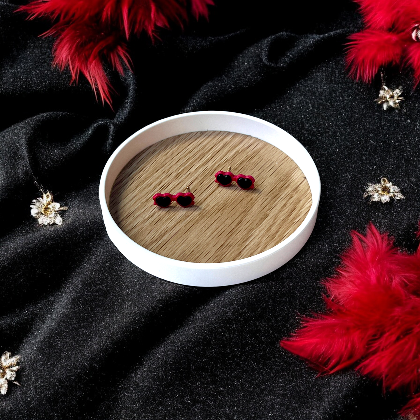 Red heart sunglasses earrings placed in a wooden tray, surrounded by sparkly black fabric, red feathers, and decorative golden accents.