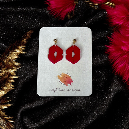 Red lips-shaped dangle earrings displayed on a Craft Love Designs card, set against black velvet fabric and vibrant red feathers.