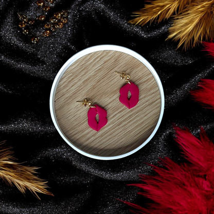 Red lips dangle earrings placed in a wooden tray, surrounded by black and gold fabric, red feathers, and decorative golden accents.