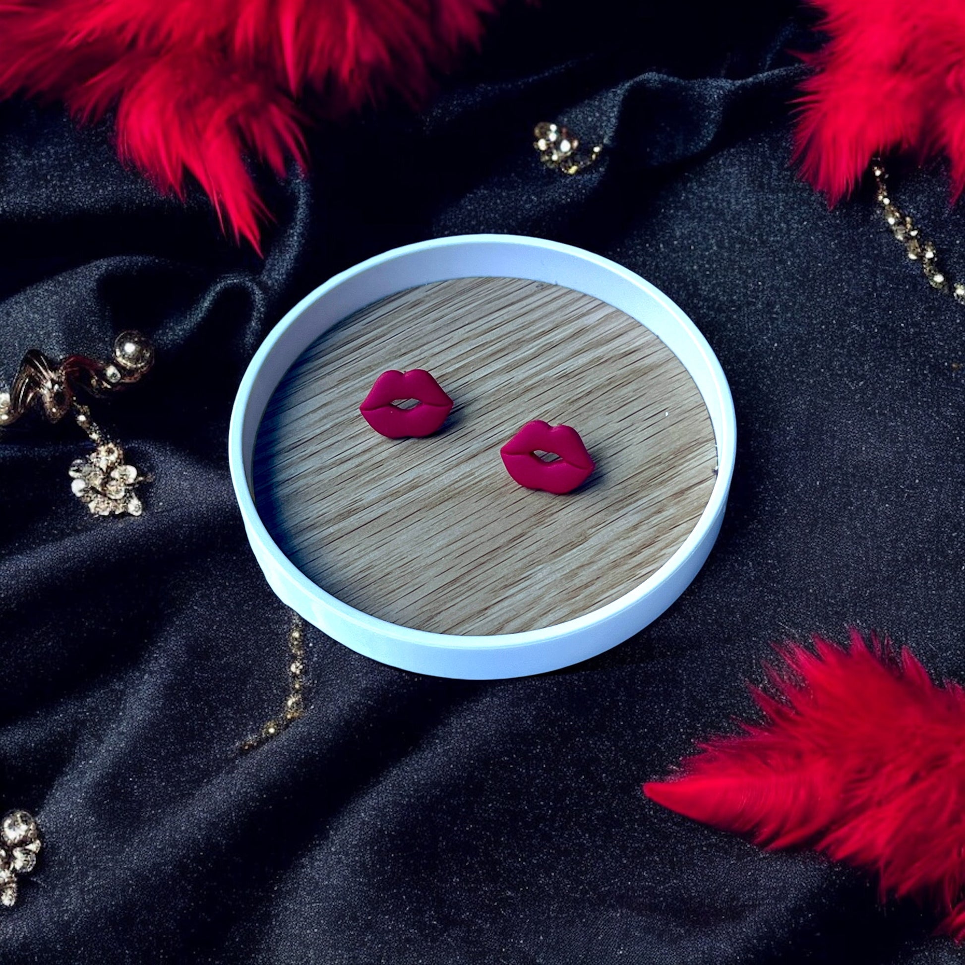 Red lips stud earrings placed on a wooden tray, surrounded by luxurious black fabric, sparkling embellishments, and vibrant red feathers.