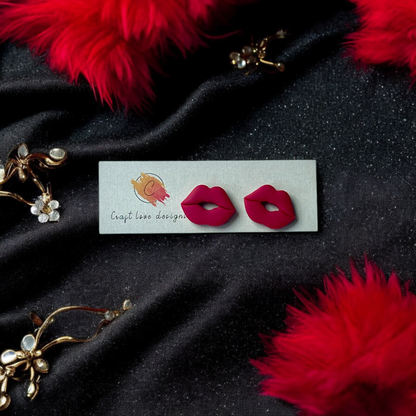 Red lips-shaped stud earrings displayed on a Craft Love Designs card, set against black velvet fabric with red feathers and gold accents.