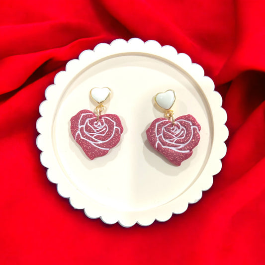 Red rose heart dangle earrings featuring sparkling glitter roses and white heart studs on a round white plate over a vibrant red background.