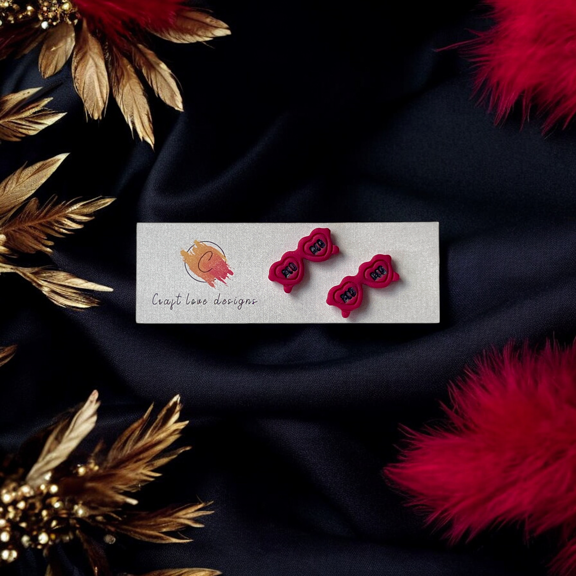 Fun red sunglasses-shaped earrings with "Red" text displayed on a Craft Love Designs card, set against luxurious black fabric and red feathers.