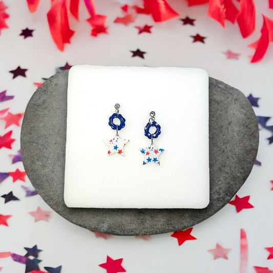 Handmade dangle earrings with red, white, and blue star patterns, paired with blue accents and rhinestone studs on a white cushion.