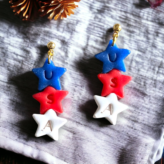 Handmade polymer clay USA star dangle earrings in red, white, and blue, with gold hardware, displayed on a textured surface.