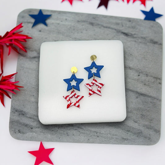 Patriotic double star dangle earrings with blue open stars and red striped stars, accented with gold studs, on a white cushion.