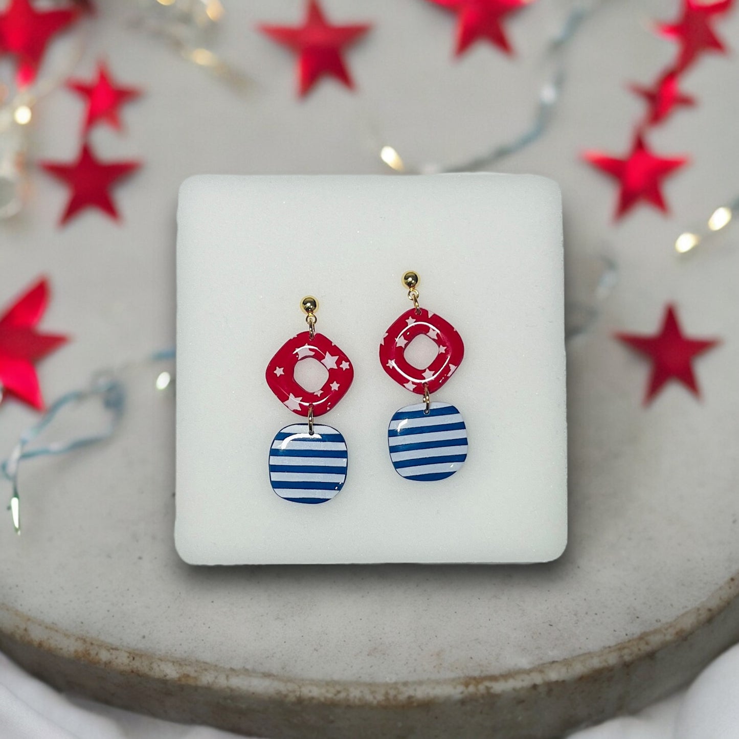 Handmade geometric 4th of July dangle earrings featuring red stars and blue stripes, displayed on a soft white cushion.