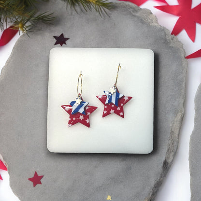 Patriotic red and blue star hoop dangle earrings with layered stars and gold accents, displayed on a soft white cushion.