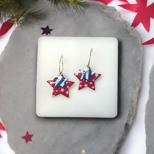 Patriotic red and blue star hoop dangle earrings with layered stars and gold accents, displayed on a soft white cushion.