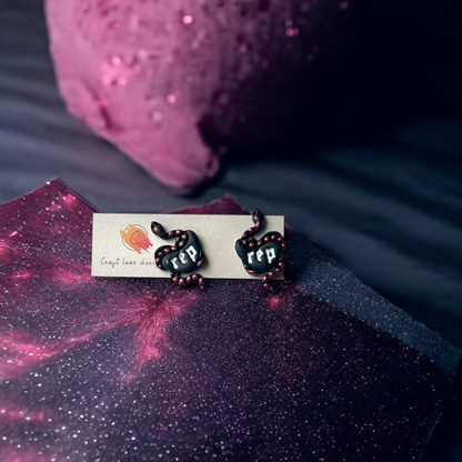 Pair of black heart earrings with “rep” logo and coiled pink-accented snakes, resting on glittery wrapping paper in a cosmic pink-hued setting.