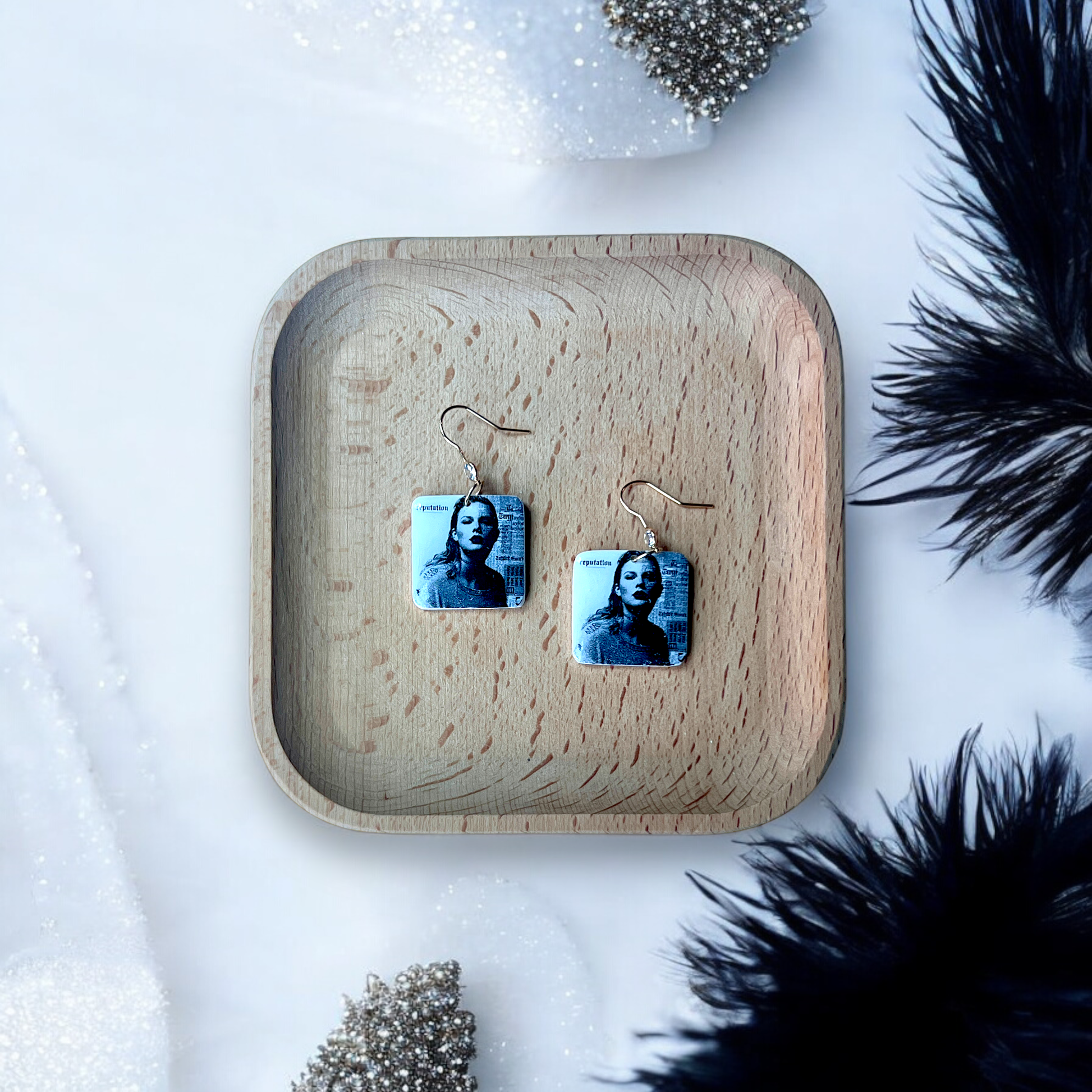 Square dangle earrings featuring a black-and-white portrait inspired by the Reputation album, displayed in a wooden tray with soft decor accents.