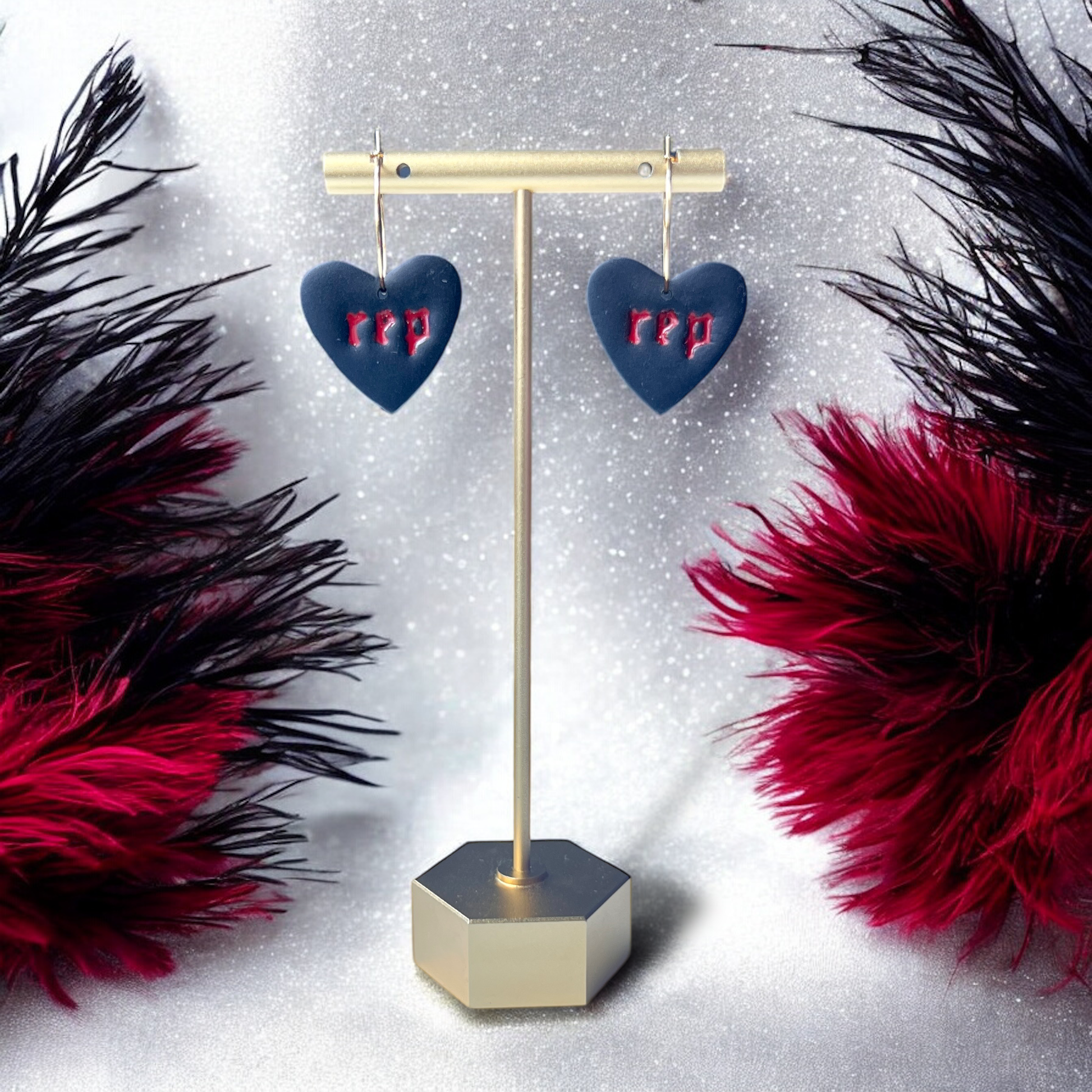 Heart-shaped back earrings featuring red "rep" text, displayed on a branded card with black and red feather accents. 
