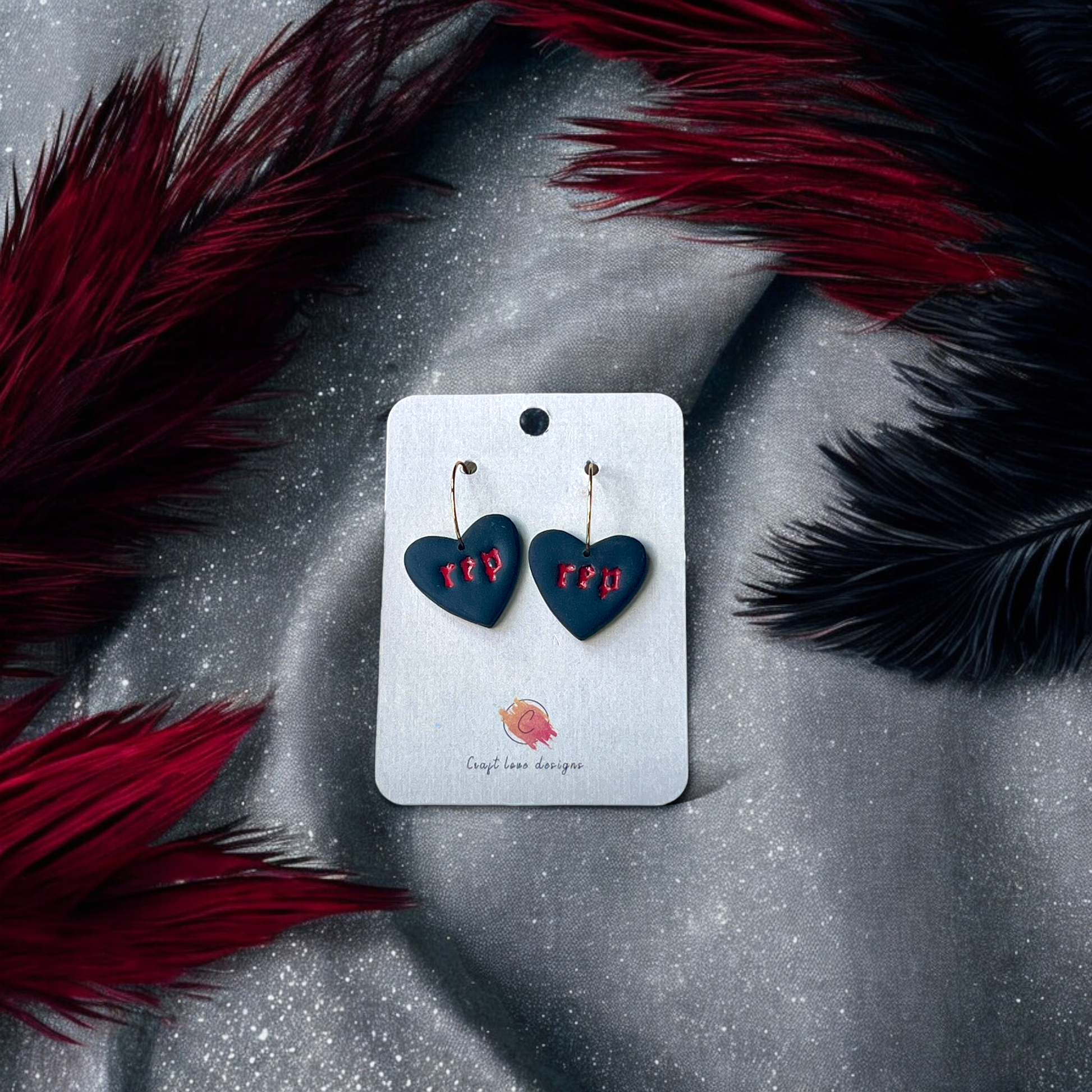 Black heart-shaped drop earrings with "rep" text in red, displayed on a gold stand with feather accents for a bold and elegant look.