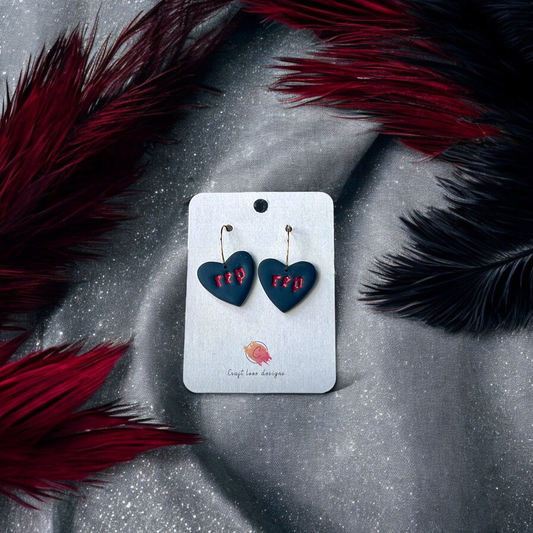 Black heart-shaped drop earrings with "rep" text in red, displayed on a gold stand with feather accents for a bold and elegant look.