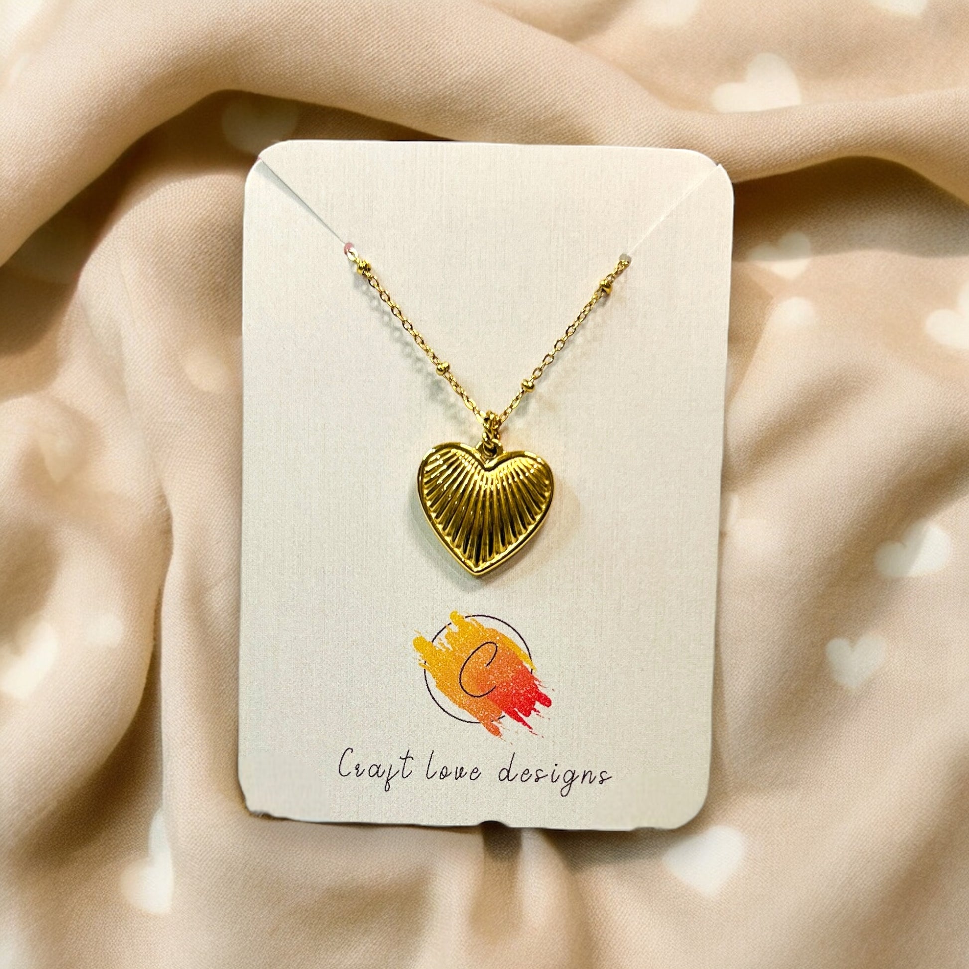 Elegant gold necklace with a ribbed heart pendant showcased on a branded card, surrounded by soft beige fabric featuring faint heart accents.